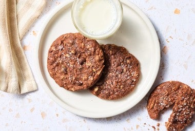 Two Chocolate Lactation Cookies on a plate with a glass of milk and another cookie on the counter next to the plate.