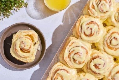 Fluffy Frosted Orange Rolls seen from above on a sheet tray with a single roll on a dark plate.