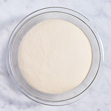 Pretzel bun dough in a clear glass bowl after rising to double size.