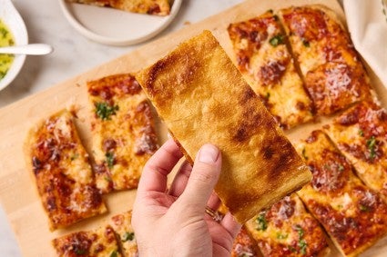 A hand holding a slice of flaky puff crust pizza upside down to show the golden and dark brown crust underneath.