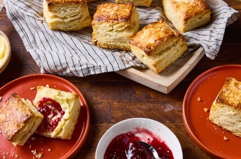 Overhead view of a sliced biscuit on a red plate and a wooden table. There is red jelly and butter on the biscuit slice.
